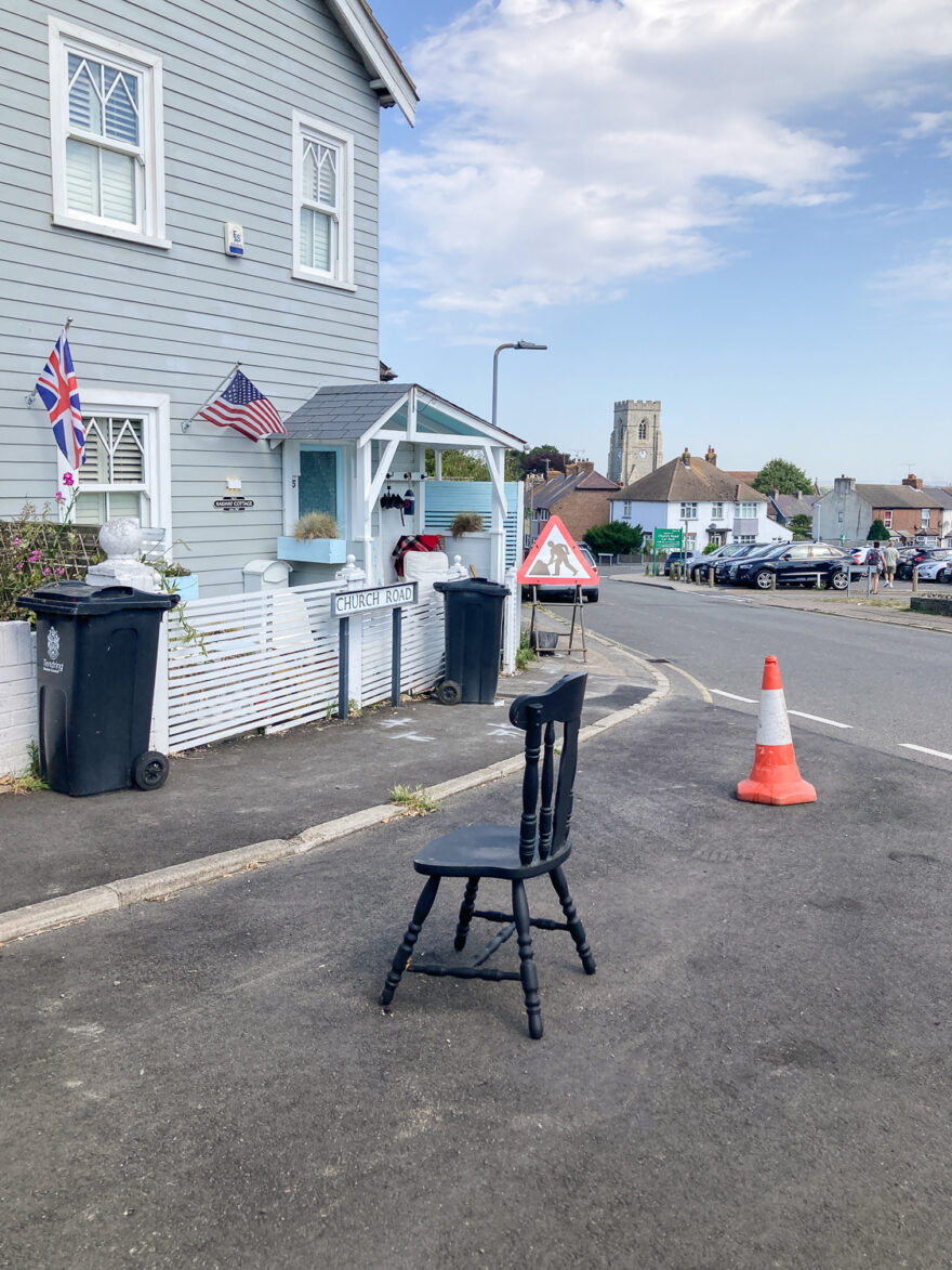 Photograph of a black wooden kitchen chair in the road outside a house, with traffic cone to the right behind. The house has a light blue wooden facade, British and US flags flying and black wheely bins. In the distance is a construction work sign, a car park and buildings including a church.