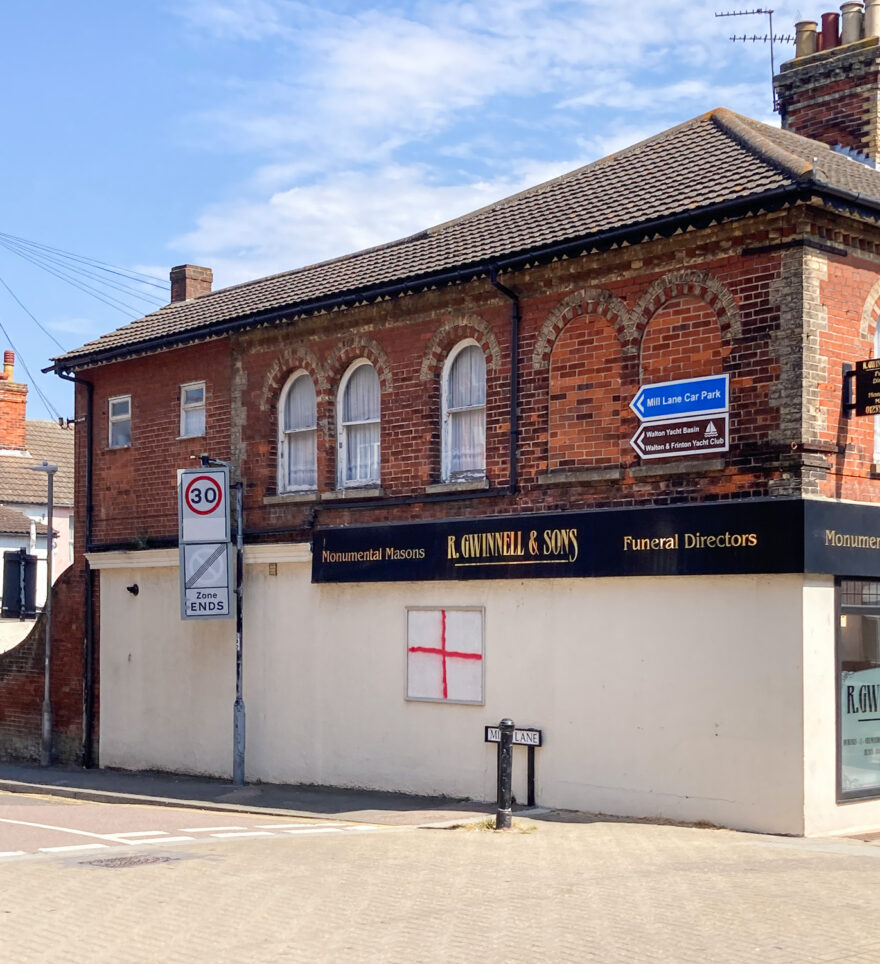 Photograph of the side of a Funeral Director's building, with an empty white noticeboard that has had a red cross sprayed on it, to resemble the England flag.