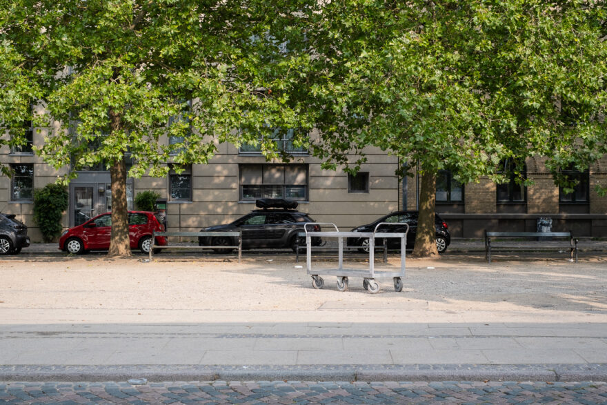 Copenhagen, Denmark, August 2024. Photograph across a street to a large metal trolley that's been left in the middle. Behind are trees, parked cars and buildings.