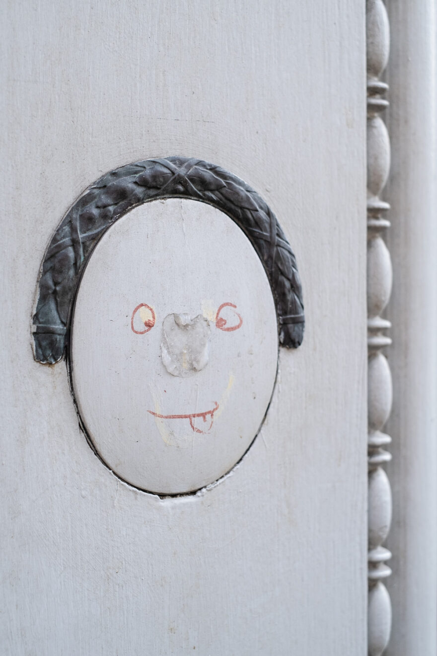 Face drawn on an ornate wall feature, Copenhagen, Denmark. Copenhagen, Denmark, August 2024. Photograph of an ornate architectural element framing the top of an oval on a wall, with eyes and mouth with tongue poking out drawn in red marker within it, so the ornate part resembles hair. To the right is an ornate bar lining the length of the frame.