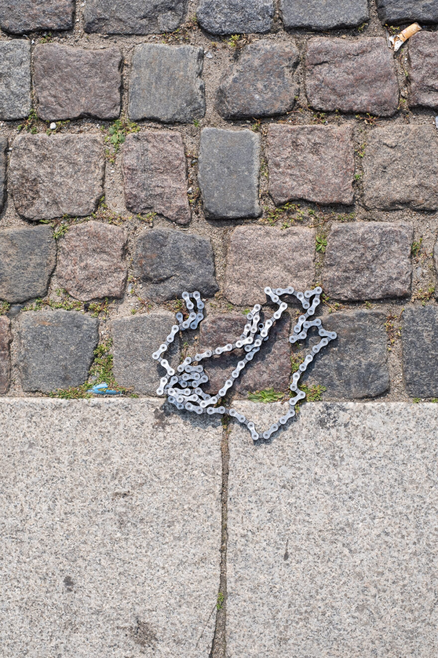 Broken bike chain on the floor. Copenhagen, Denmark, August 2024. Photograph looking down at a broken bike chain lying on a cobbled road, with larger concrete slabs at the bottom of the frame.