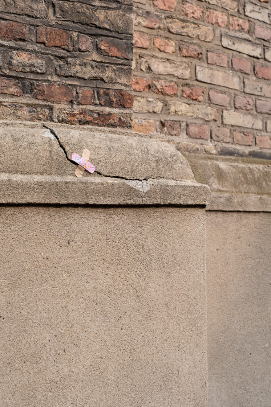 Copenhagen, Denmark, August 2024. Photograph of a wall, brick at the top and concrete in the lower third, with a cross of medical plasters (one beige, one pink unicorn) stuck over a cracked part of the wall.