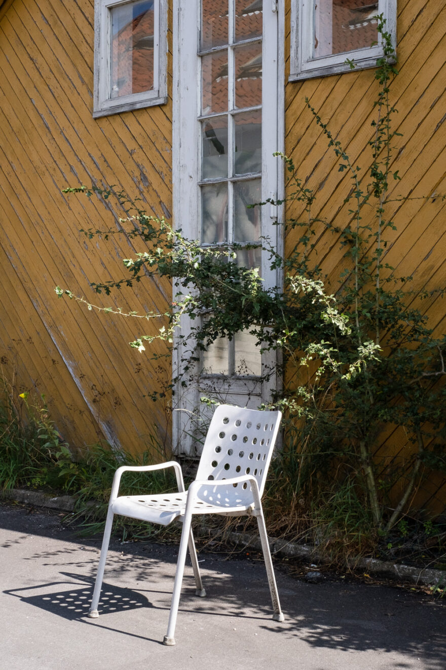 Christiania, Copenhagen, Denmark, August 2024. Photograph of a white plastic chair with circular holes evenly distributed across the surface, in the sunlight at the side of the road. Behind is a building with diagonal yellow planks across the walls, with white-framed windows and green plants along the bottom and growing up and arching over the chair.