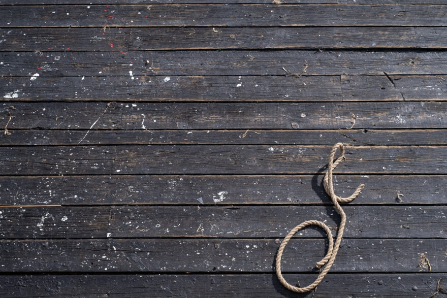 Copenhagen, Denmark, August 2024. Photograph looking down at a wooden floorboard floor, painted black and worn, with paint, dirt and glass across it. In the lower right of the frame is a length rope, curled so it looks a bit like an ornate J.