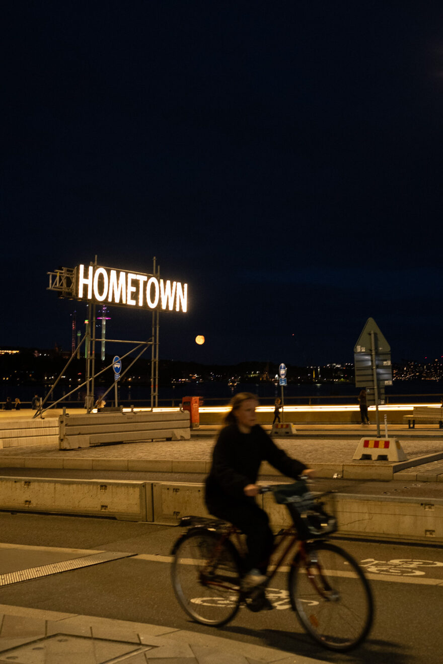 Stockholm, Sweden, August 2024. Photograph taken at night of a lit up sign saying 'HOMETOWN', on scafolding on a bridge with lots of signs and concrete blocks around as it's under construction. In the distance the moon can be seen bright orange, almost full. In the foreground a person on a bike is cycling past, blurred with the motion.