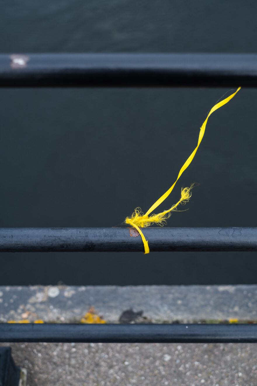 Stockholm, Sweden, August 2024. Photograph of a frayed length of yellow ribbon tied to a metal railing, blowing in the wind, with another metal railing above and below. In the background is grey water of a river and concrete floor at the bottom of the frame.