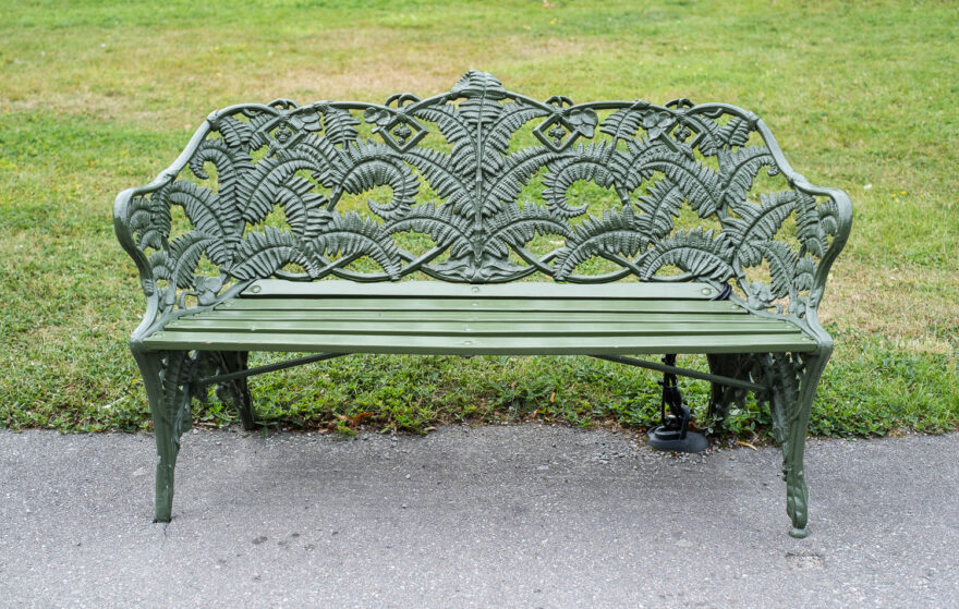 Victorian Coalbrookdale fern bench, Stockholm, Sweden. Photograph of a metal and wood bench, painted green, with an ornate fern pattern on the back and sides. Below is a tarmac path and there's a grass lawn behind.