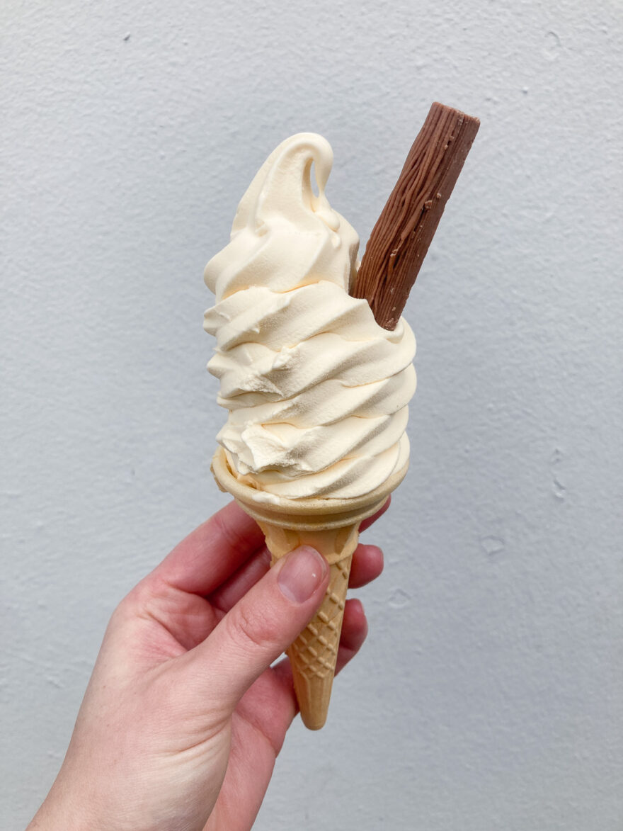 Photograph of a white hand holding a 99 whipped ice cream in a cornet, with a chocolate flake poking out to the right and a light grey wall behind.