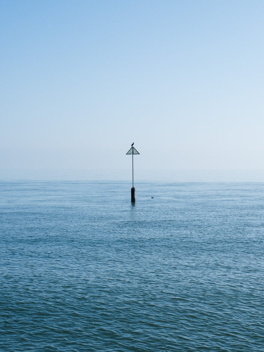 Photograph of the sea with a pole sticking out with a triangular wooden shape on the top, upon which a bird sits. A hazy blue sky is behind.