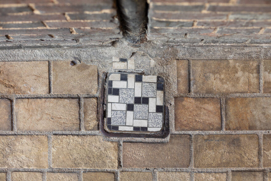 Photograph looking down at a brick floor, with a small utility cover in the centre, with an 80s style small grey, white and black tile patternon it. A brick wall with a black pipe is visible out of focus at the top of the frame.