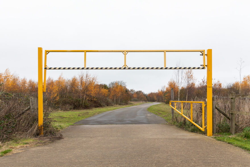 Colchester, UK, November 2024. Photograph of a metal entrance barrier painted yellow, with the gate open to the right. A concrete path goes from the foreground through the gate and winds to the right into the distance, with grass and trees either side, many of which have golden leaves. The sky is white with clouds.