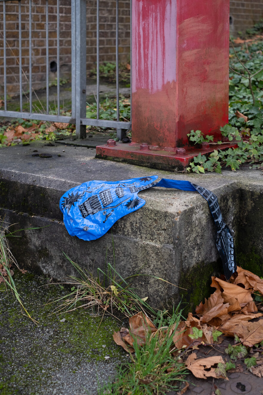 Photograph of a blue plastic inflatable guitar, deflated and resting on a raised concrete area at the side of a path, with a red metal upright coming from the top and a grey metal fence to the top left. The area around is overgrown with plants, moss and dry fallen leaves.