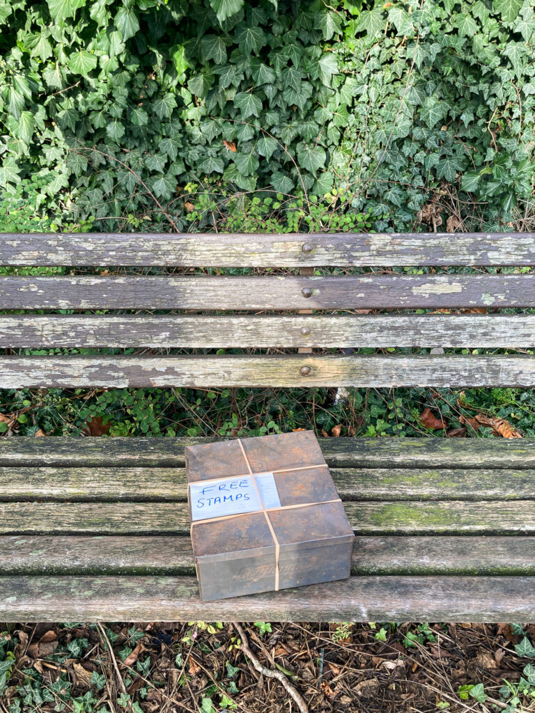 Photograph of an old metal box held together with rubber hands, with a handwritten label saying 'FREE STAMPS' on the top. The box is on a worn wooden bench, with foliage behind.