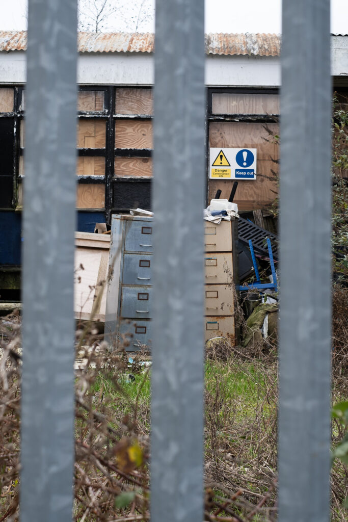 Photograph looking through 3 metal bars in a fence, towards a boarded up building with rusty old filing cabinets in front, and plants in the foreground.