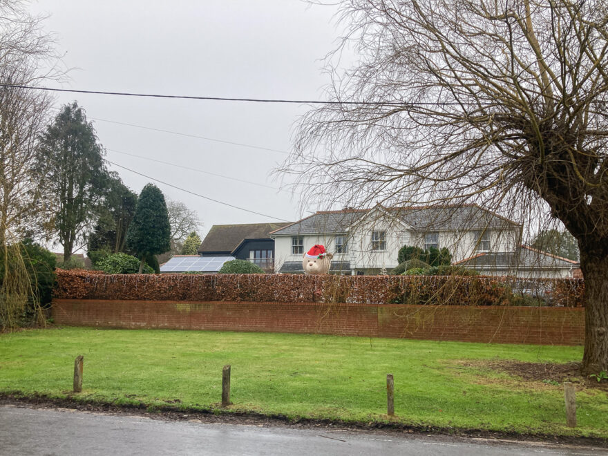 Photograph of a large house behind a brick wall, with a giant teddy with Christmas hat peering over the top. In front of the wall is a grassy area, with fence poles and a part of road visible in the foreground, with trees either side.