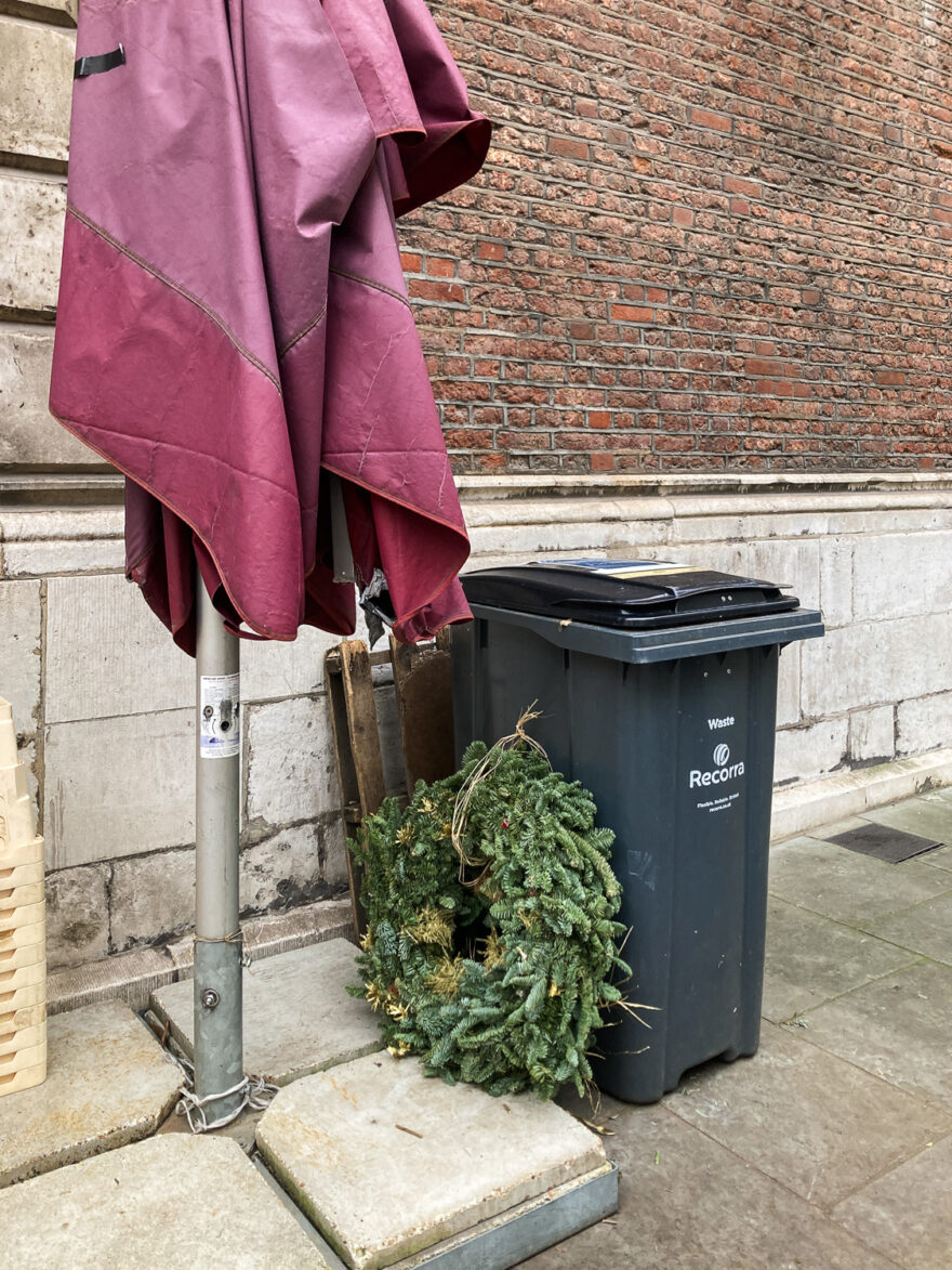 Photograph of a black wheelie bin with a large green Christmas wreath leaning against it. To the left is the bottom portion of a parasol, and a brick building is visible behind.