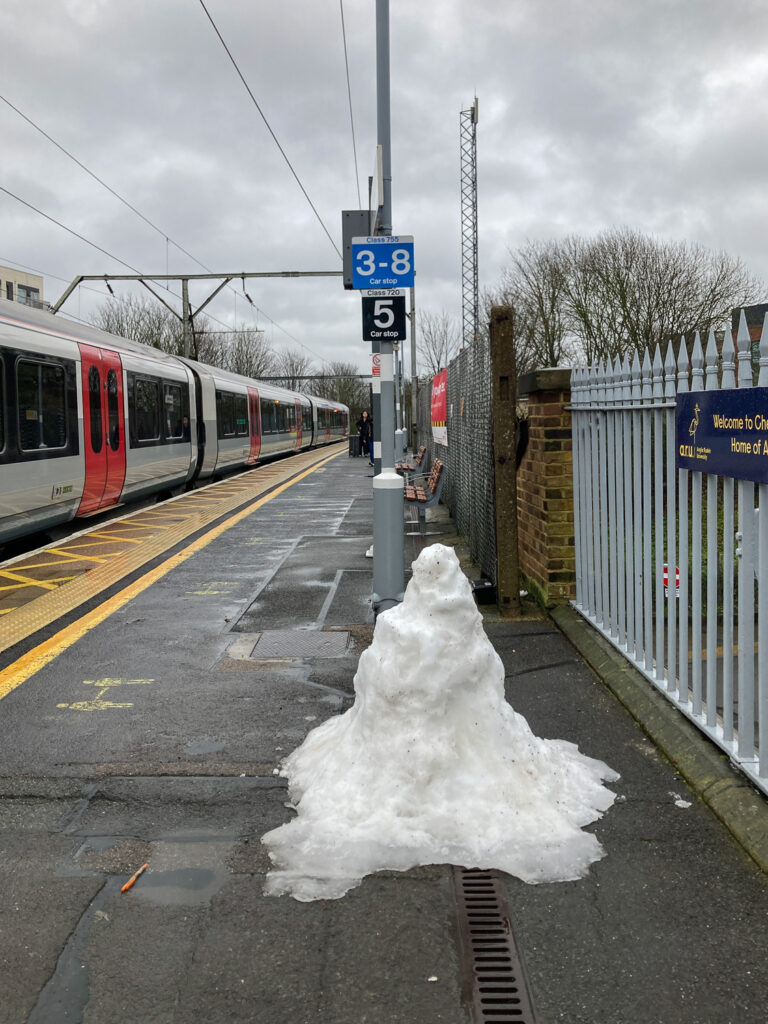 Photograph of a pile of snow (a melted snoman), on the platform at Chelmsford railway station, with a train visible to the left, posts and signs behind in the centre of the frame and grey metal fence to the right of the image.