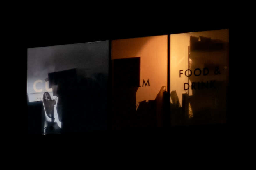 Photograph of windows in a building at night, lit from inside. To the left the silhouette of a desk fan is visible with boxes around it, through a grey window with 'CURZON' visible very faintly and to the right two orange windows with 'FILM, FOOD & DRINK' and silhouettes of boxes and shelving can be seen.