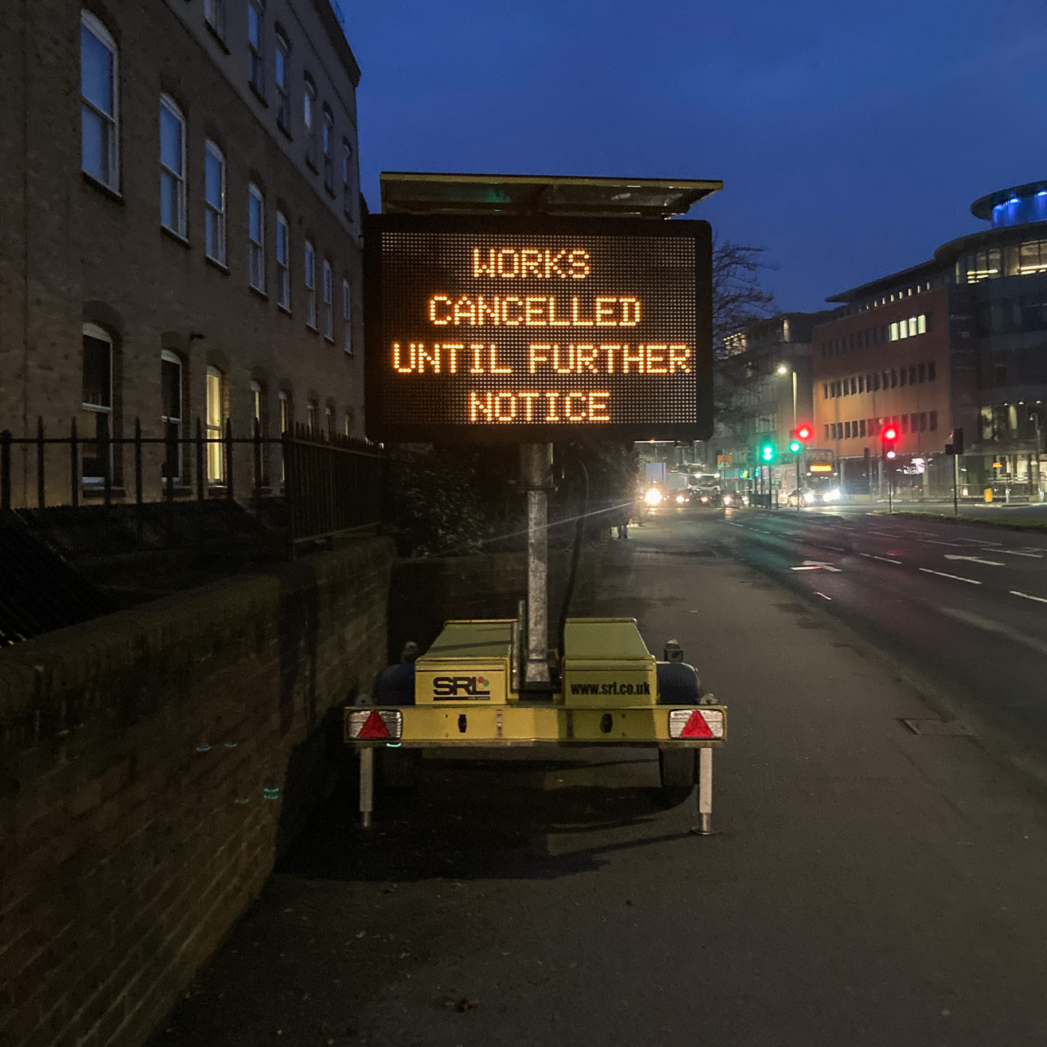 Photograph of a portable constuction notice with 'WORKS CANCELLED UNTIL FURTHER NOTICE' in orange lights on black, on a pavement with wall and buildings to the left of the frame and road with lights, traffic and buildings in the distance. The sky above is dark blue as it's early in the morning.