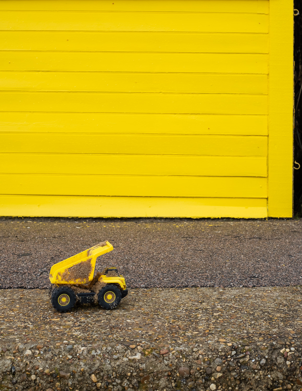 Photograph of a small yellow plastic toy pickup truck covered in sand, on a concrete floor with a bright yellow beach hut behind.