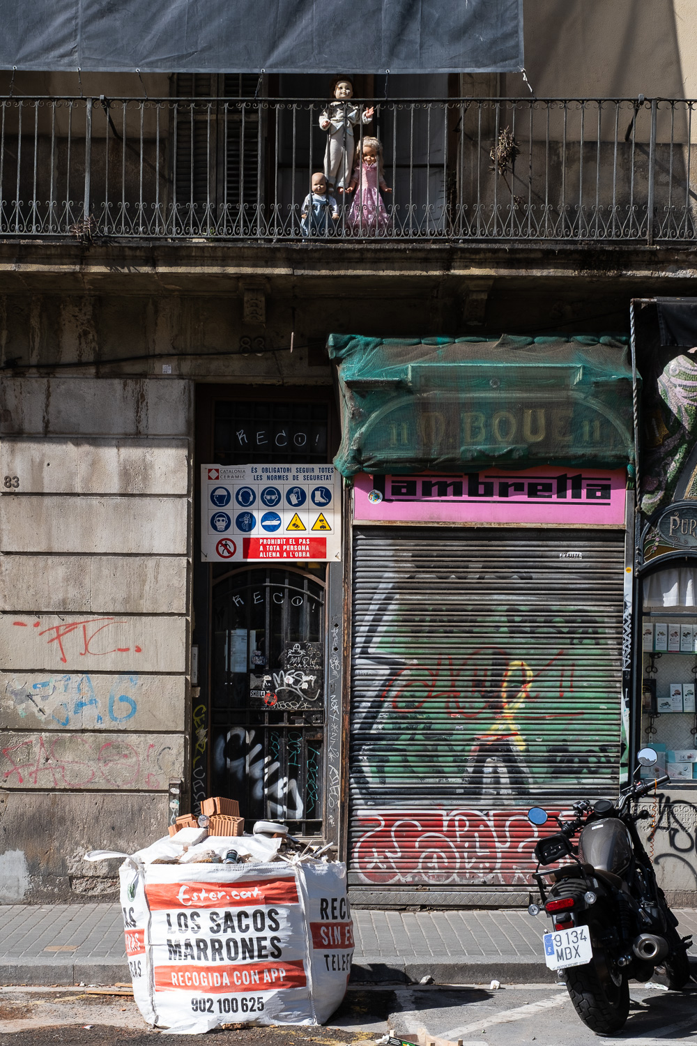 Photograph of a balcony with three dolls stood up, looking a bit creepy. Below is a building underconstruction, with shutter saying 'Lambretta', grafitteed shutter underneath, sack of construction materials and motorbike on the road in front.