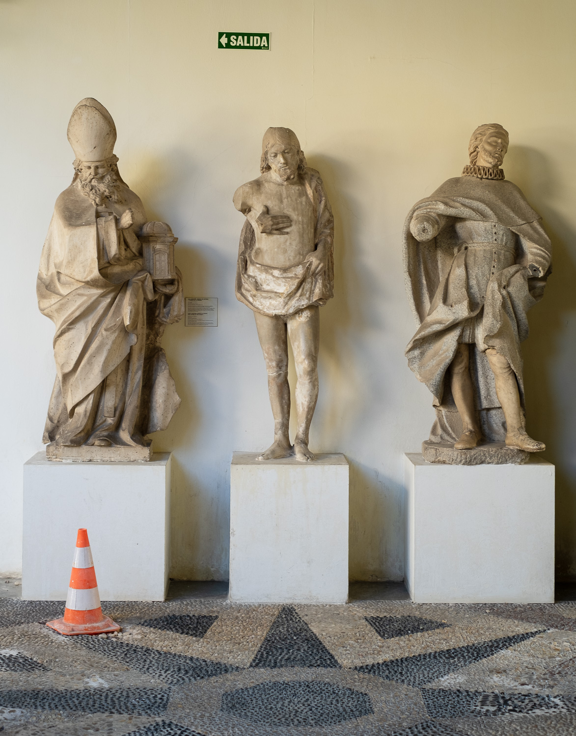 Photograph of three statues on white plinths, with an orange and white plastic cone to the left, where the statue behind it appears to be looking at it. On the wall is a green sign pointing left and saying 'SALIDA'.