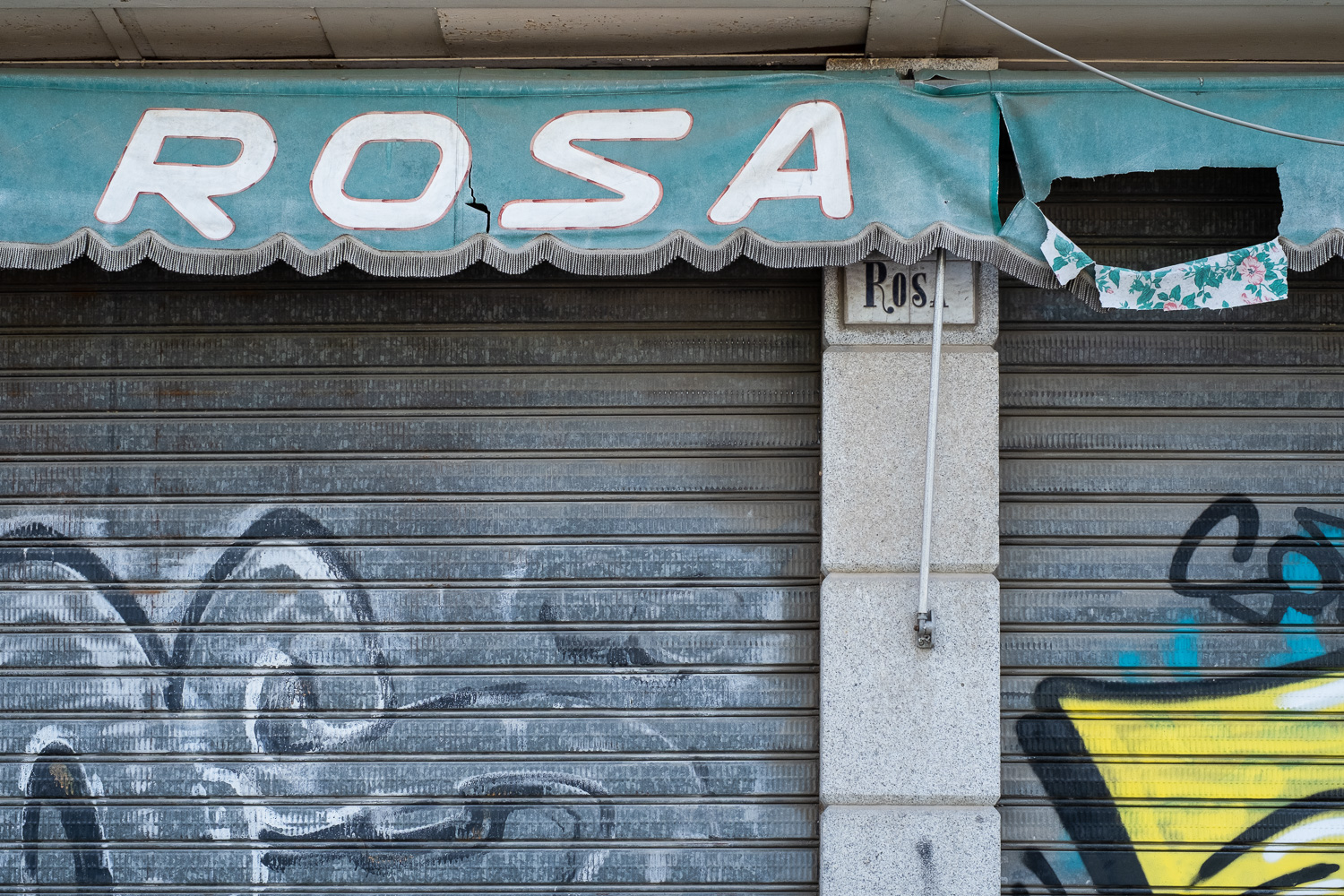 Photograph of a shuttered shop front with a canopy above saying 'ROSA'. To the right the canopy is torn and flapped over, showing a floral rose pattern on the reverse of the fabric.