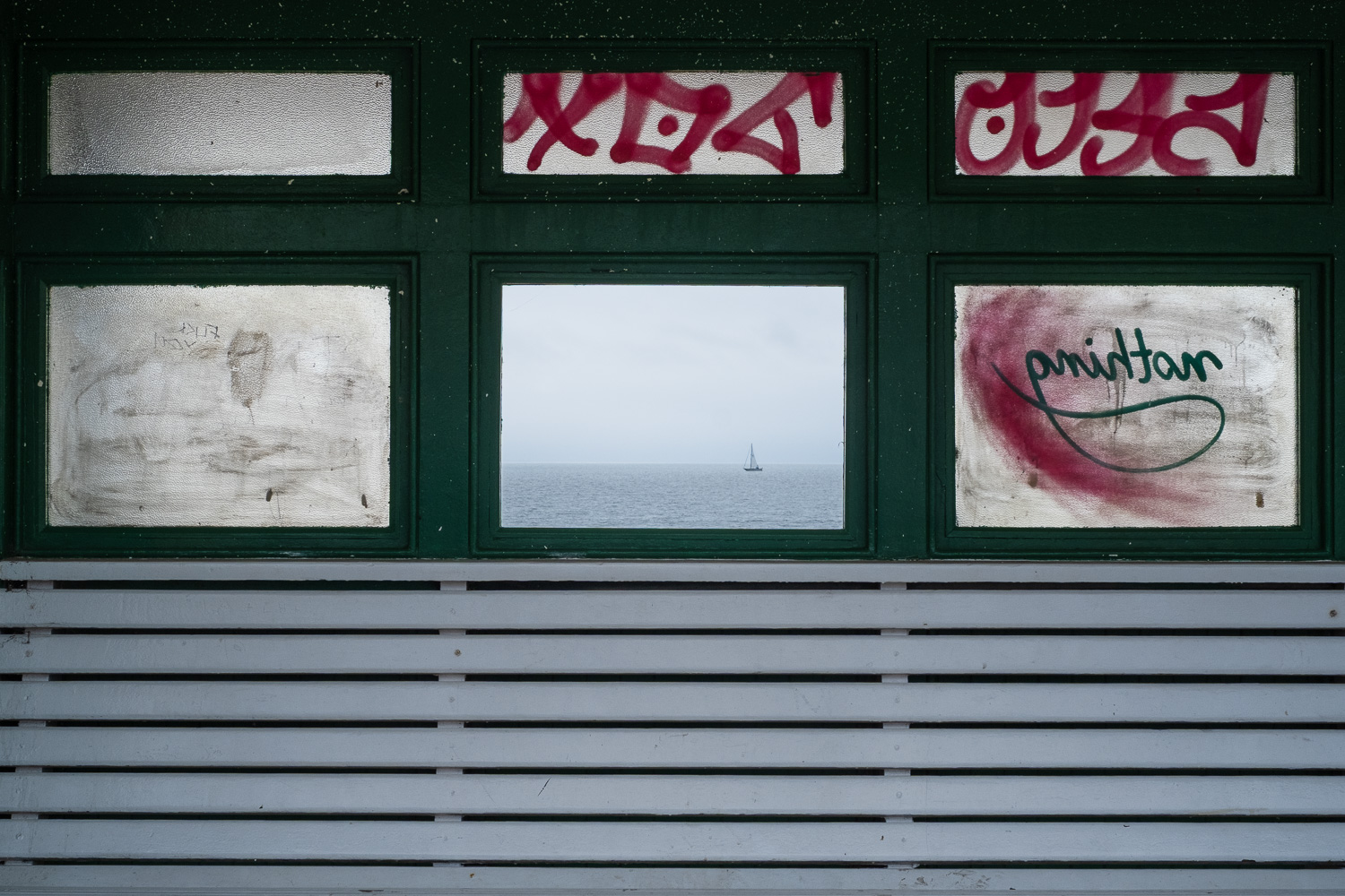 Sea view. Photograph through a covered bench area on the seafront. The lower centre pane of glass is missing and the sea and a boat can be seen through it. The surrounding windows are dirty and have graffiti on them. The wood inbetween is painted dark blue and the white wooden bench can be seen in the lower third of the frame.
