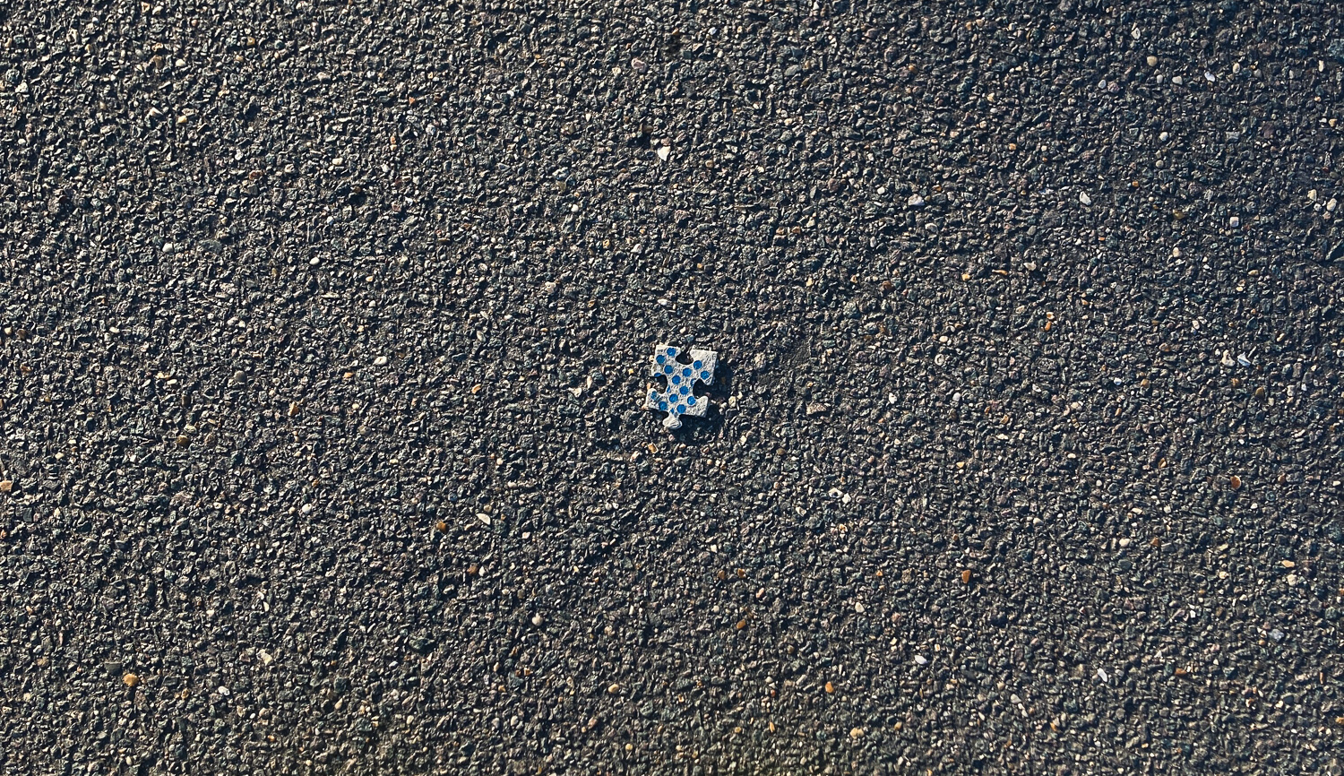 Photograph of a grey puzzle piece with blue dots on it, on a tarmac pavement.