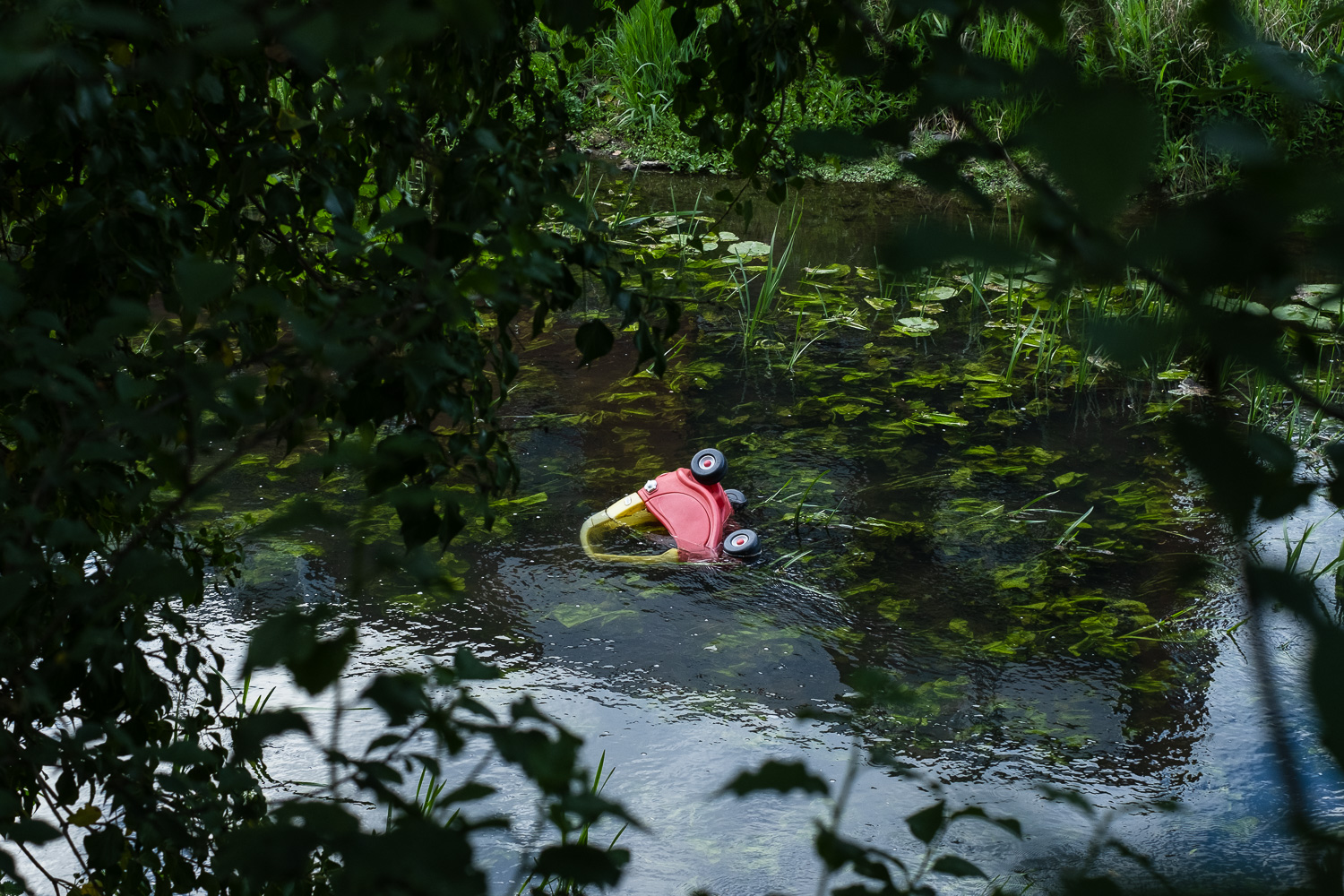 May 2025, Colchester, UK. Photograph of a red and yellow Little Tikes Cosy Coupe plastic toy car in a river, half in half out of the water, with river plants all around it. The photograph is taken from the river bank, looking through trees, which can be seen out of focus in the foreground around the frame.