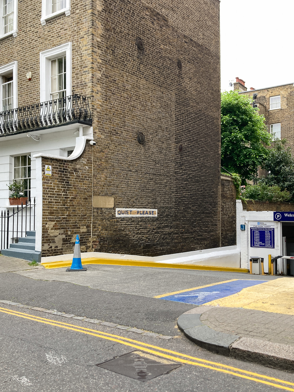 Photograph of the entrance slope to a parking area, with 'QUIET PLEASE' painted on the brick wall side of a three-storey house. There is a blue traffic cone at the top of the slope and yellow and blue painted areas on the ground.