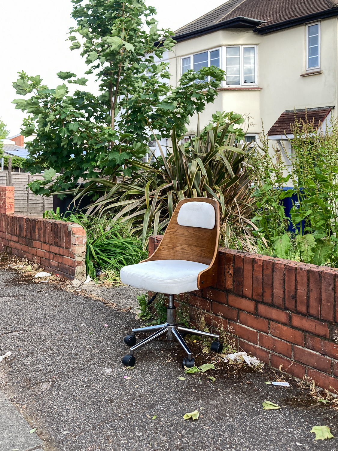 Photograph of a wood-backed office chair on a pavement with a red brick wall, garden and house behind.