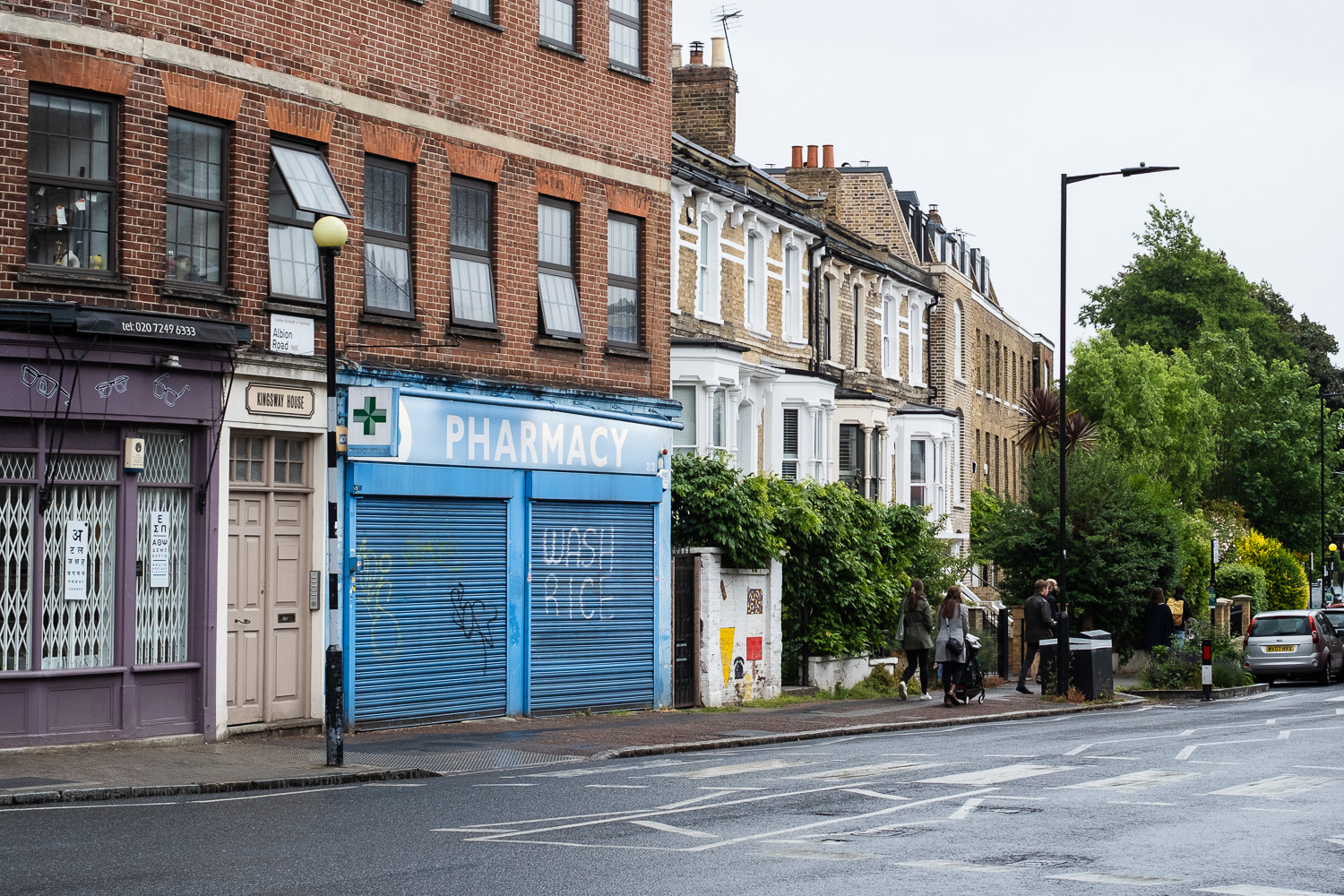 Photograph of a London street, with a Pharmacy in the centre, with its blue shutter pulled down and 'WASH RICE' written in white paint. There is a pedestrian crossing in front and houses and plants to the right of the frame.