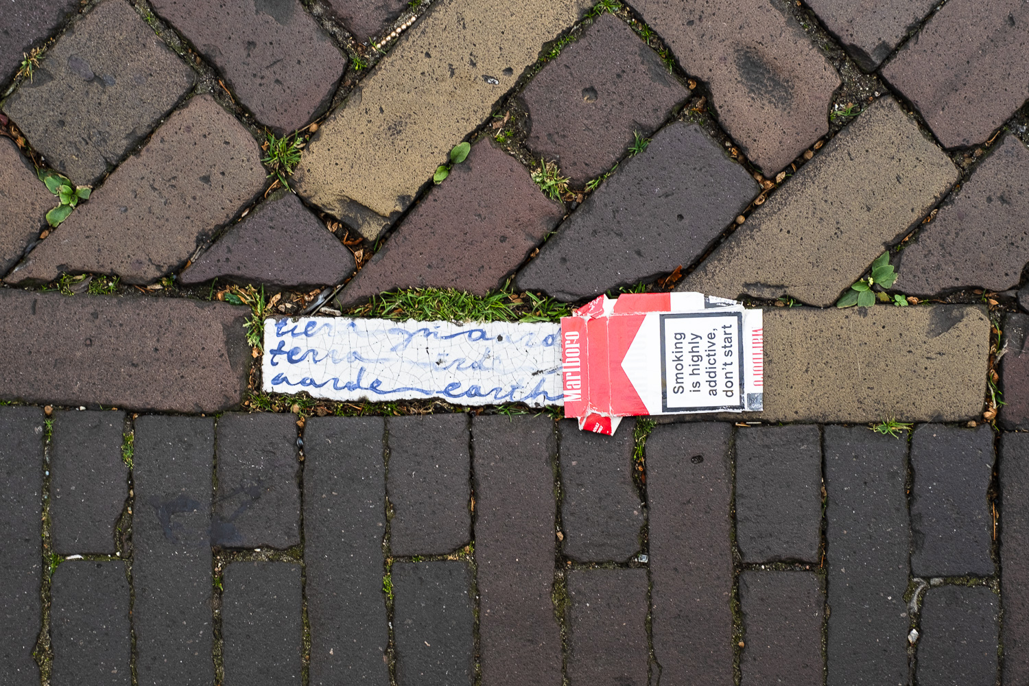 Photograph looking down on a brick paved pavement with a ceramic tile with handwritten words including 'terra... earth...', in delft blue on whte, with a discarded Marlboro cigarette packet squashed next to it, lined up so it looks as though the tile is coming out of the packet. The packet has a warning saying 'Smoking is highly addictive, don't start' on it.