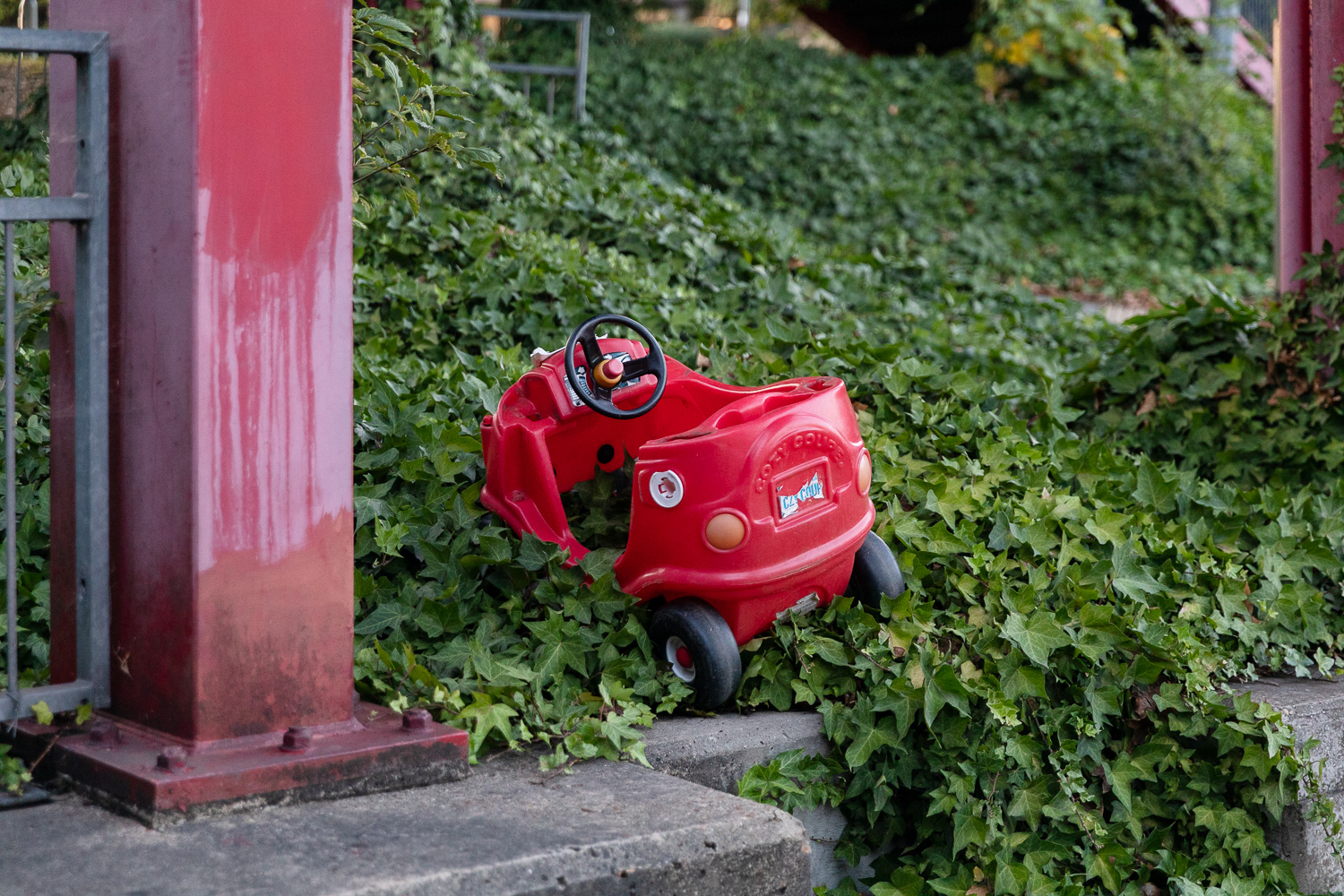 Photograph of a broken Little Tikes red plastic toy car without roof or door, dumped in a verge of overgrown ivy, between red pillars, with a low concrete wall in the foreground.