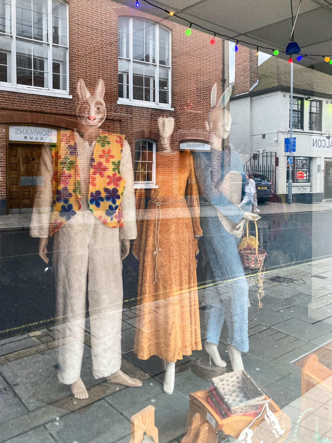 Photograph of three mannequins through a window, with reflection of a street with buildings, pavement and a road on the glass in front. The mannequins are wearing 70s style clothes with rabbit ears and masks.