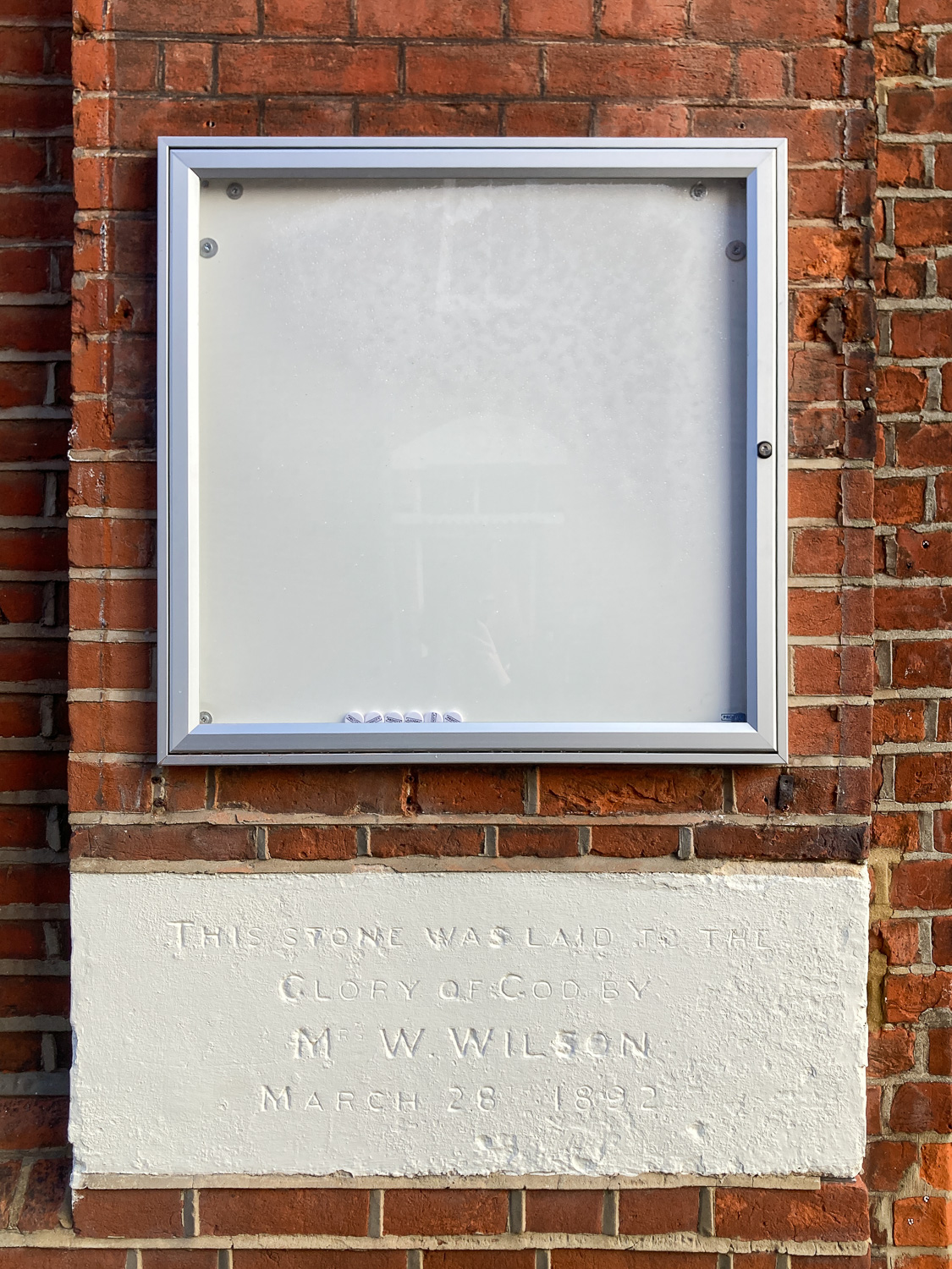 Photograph of an empty noticeboard with metal frame, magnets at the bottom of the frame, mounted on a brick wall, with a faded engraved white placque below.