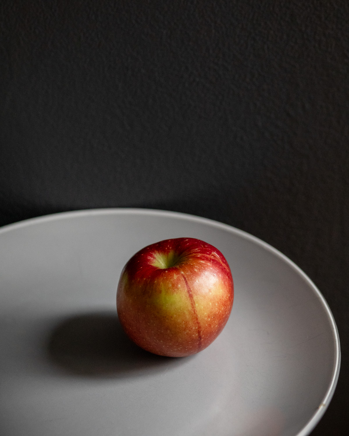 Photograph of a red and green apple on a grey plate, with a dark grey wall behind. The apple has a dark red stripe going from top to bottom.