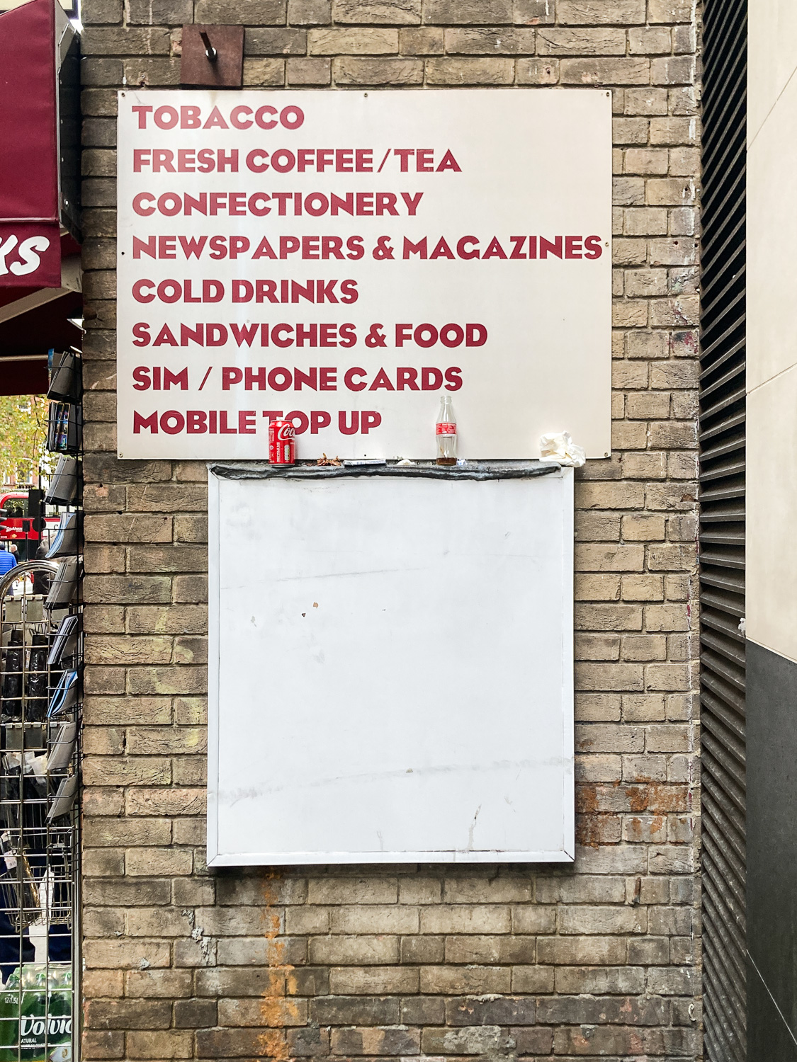 Knightsbridge, London, UK, December 2025. Photograph of an empty white sign on a cream brick wall, with a sign above that reads 'TOBACCO, FRESH COFFEE/TEA, CONFECTIONERY, NEWSPAPERS & MAGAZINES, COLD DRINKS, SANDWICHES & FOOD, SIM / PHONE CARDS, MOBILE TOP UP', and an empty Coke can and glass bottle balanced between them both. To the left a rack of postcards can just be seen along the wall.