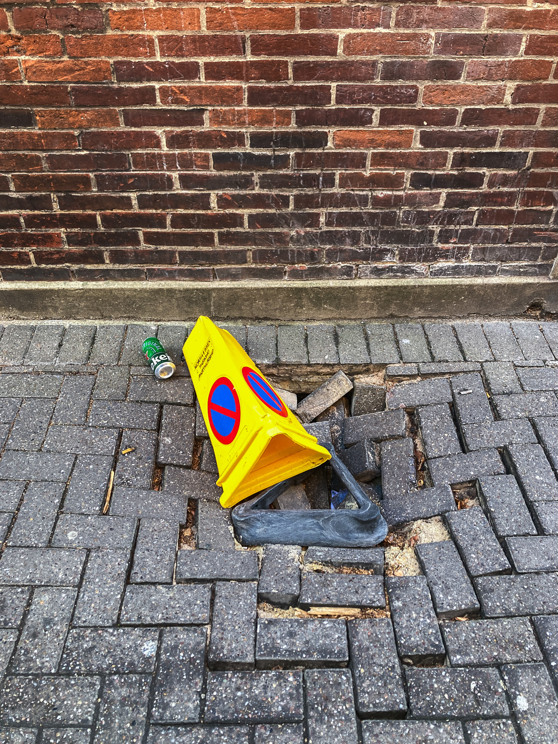 Photograph of a broken yellow plastic traffic cone with 'NORTH ESSEX PARKING PARTNERSHIP' written on it and a red and blue circular 'no entry' symbol, lying on an area of brick paving that appears to have sunk into the ground. There's a can of Heineken to the left of the cone and a brick wall across the top portion of the frame.