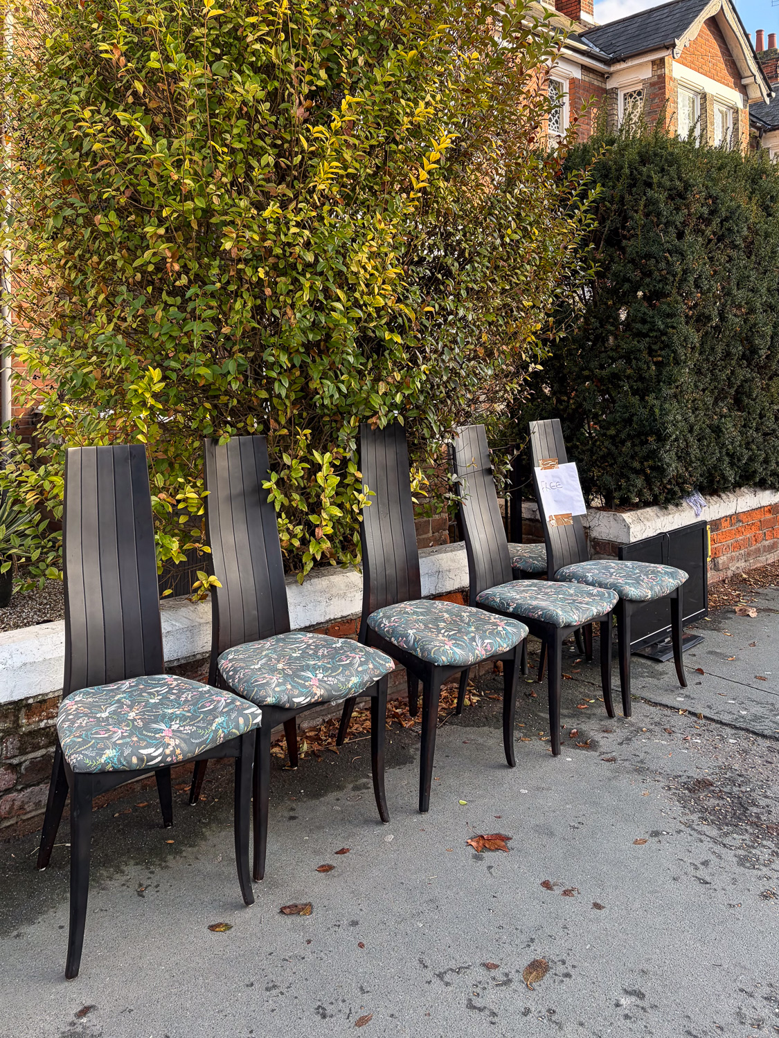 Photograph of 5 black dining chairs with floral upholstered seats, lined up against a low brick wall on a tarmac pavement. There are bushes behind the chairs and a sign saying 'FREE' is attached to the furthest chair with brown tape. A small monitor or TV screen can also just be seen at the right of the chairs.