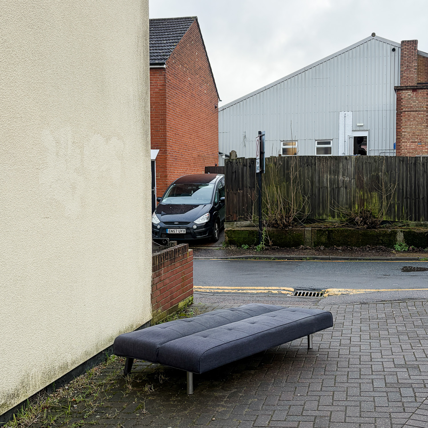 Photograph of a long dark blue upholtered seat (like a long footstool or a backless sofa) on a wide pavement, with buildings and a van around it.