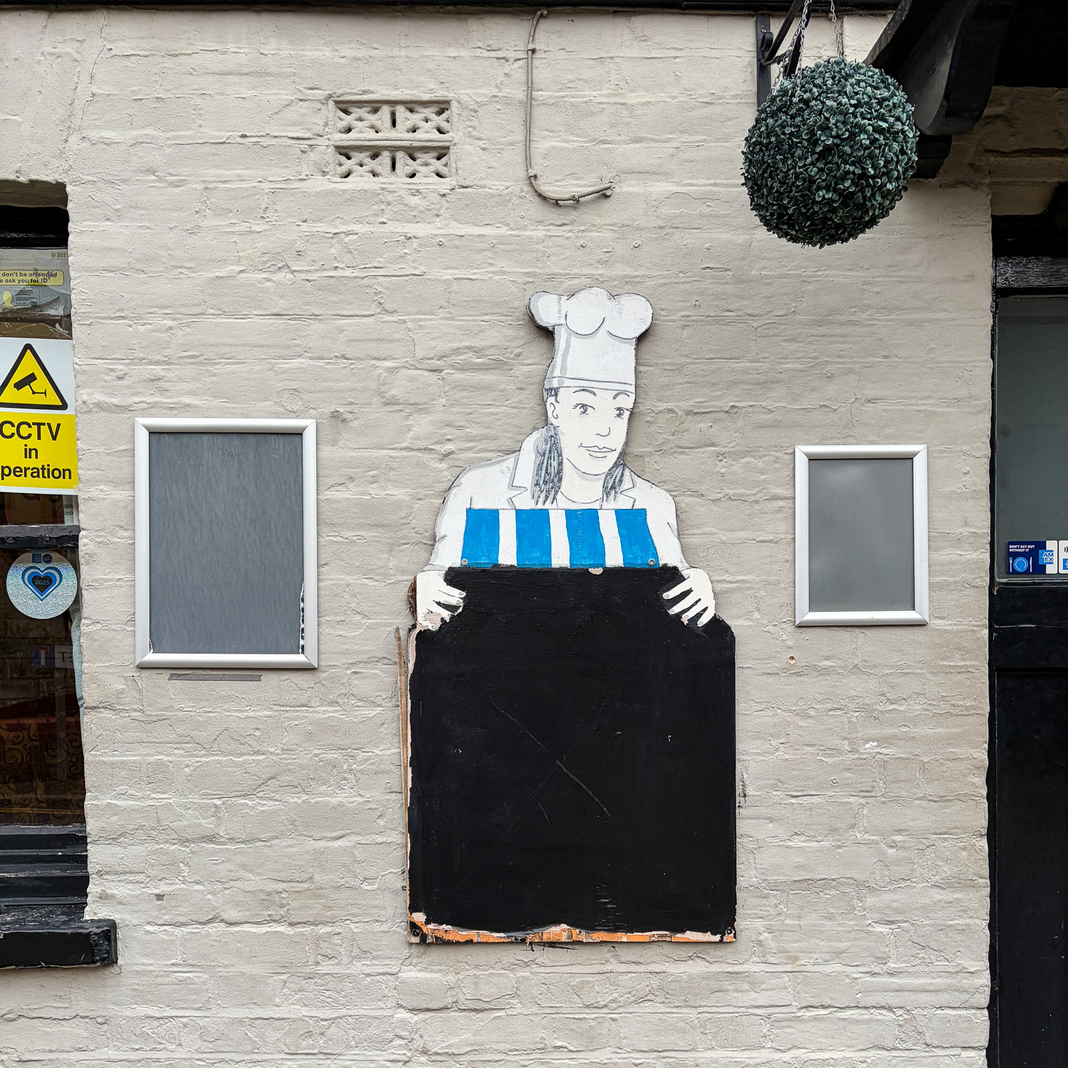 Photograph of a wall with two silver-edged empty noticeboards at either side and an empty chalkboard with an illustration of a chef holding on to it above.