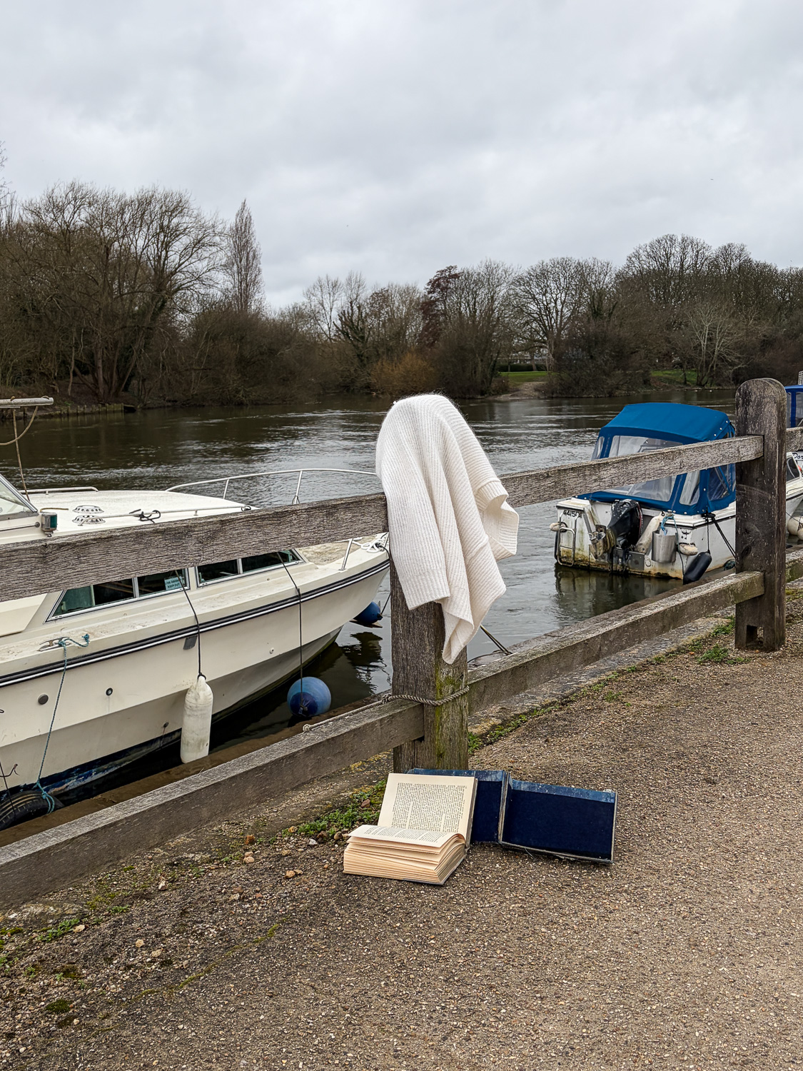 Photograph of a fluffy white cardigan on a wooden fence post, with a large hardback book on the gravel path below. Behind the fence is the River Thames, with two boats moored, and trees line the horizon beyond.