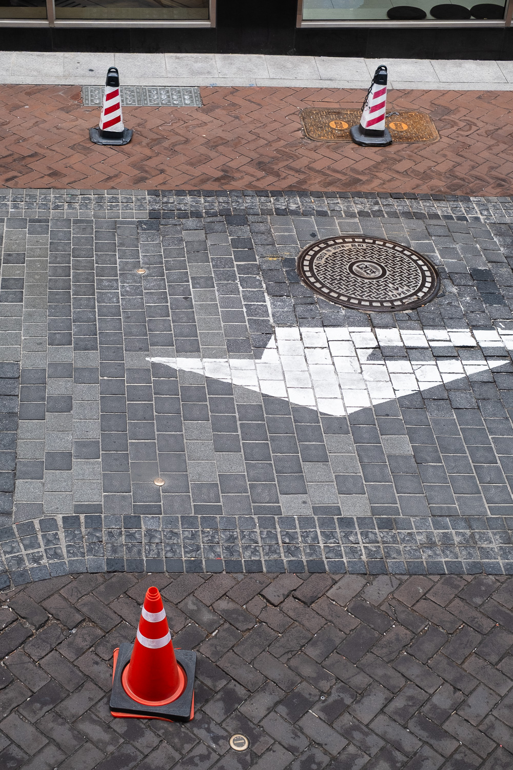 Photograph looking down at a road, with a large white arrow painted in the centre, 2 red and white striped traffic cones at the top of the frame and an orange and white striped cone at the bottom.