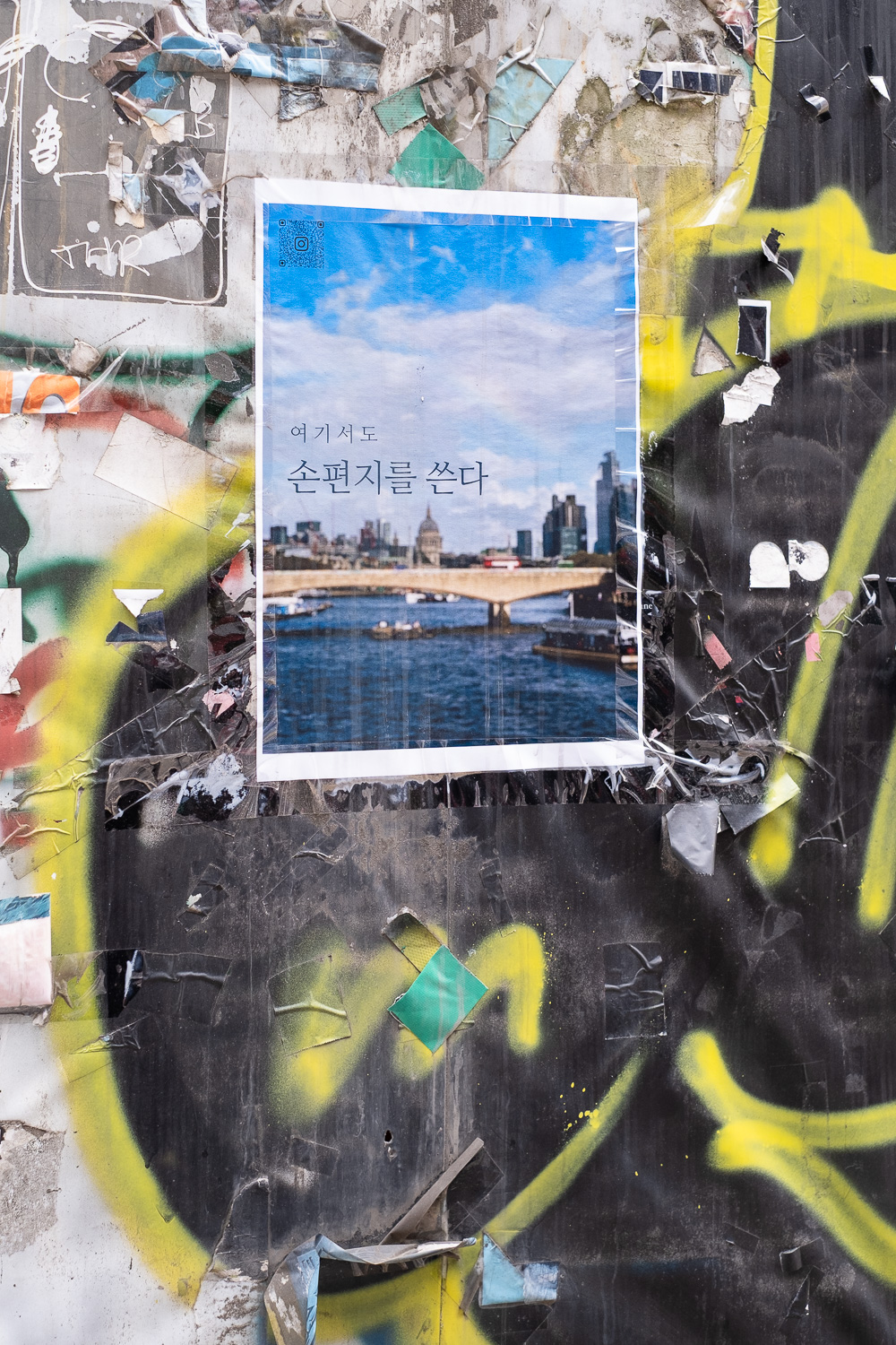 Photograph of a flyposted poster with a picture of a red bus on a bridge in London, with St Paul's in the background, stuck on a wall that has remnants of other posters and grafitti on it.
