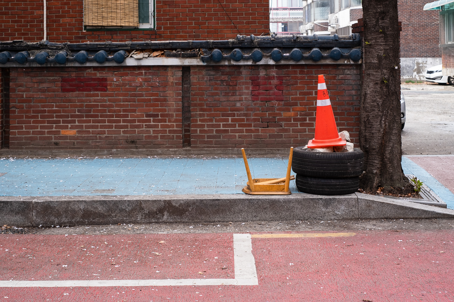 Photograph of part of a broken wooden chair and an orange traffic cone on top of two car tyres at the side of a road. The surface of the pavement is painted light blue and the road is painted red. Behind is a brick wall and there is a tree to the right.