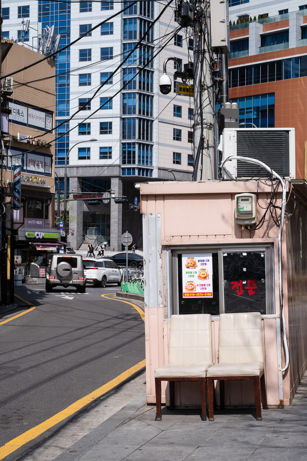 Photograph of two cream dining chairs in front of a light pink metal cabin at the side of the road. In the background are tall buildings and some cars, there is a paved pavement in the foreground and the road has single yellow line painted along the edge.