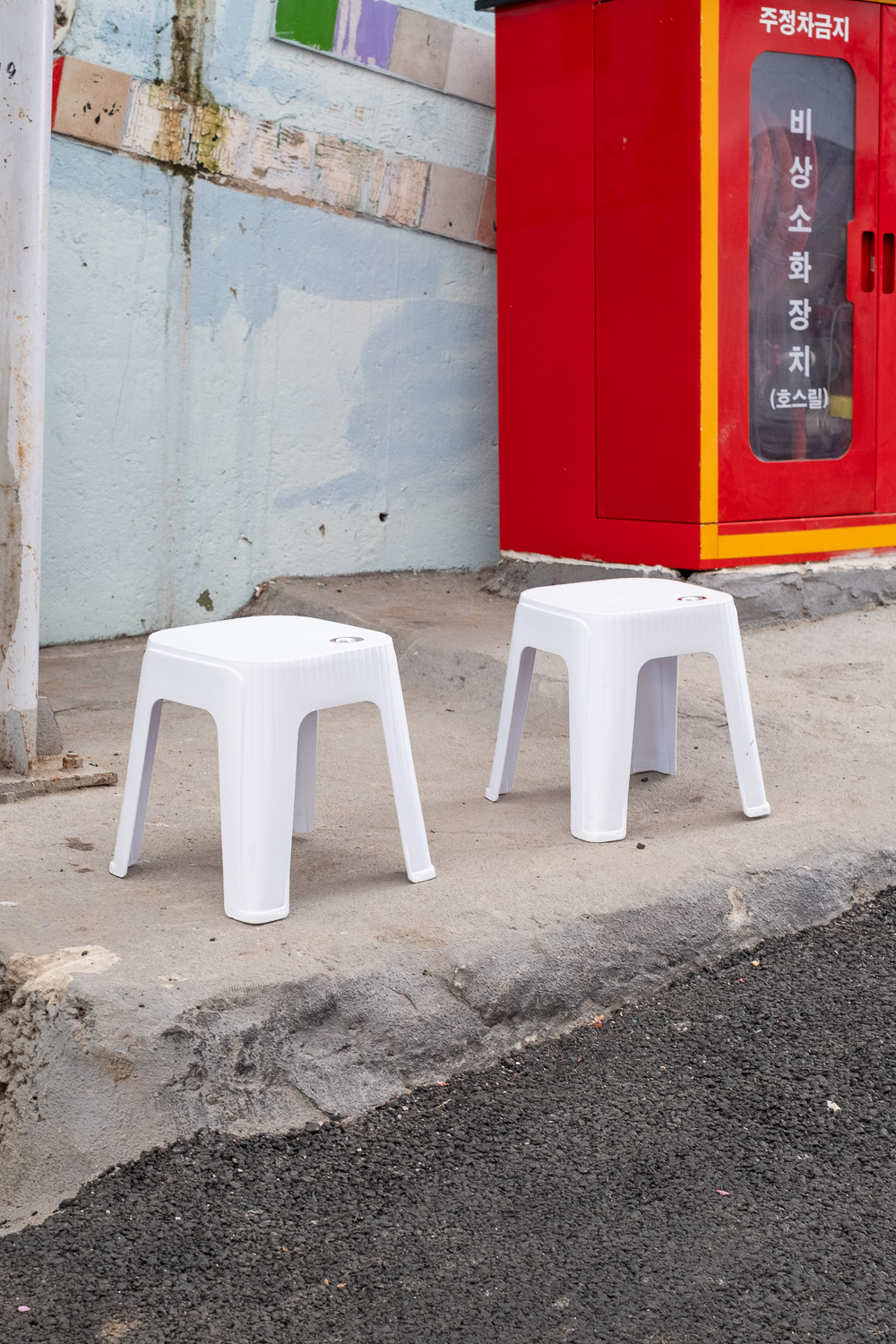 Photograph of two white plastic stools at the side of a road. A tarmace road is visible in the bottom right of the frame and behind is a blue/grey wall with a bright red fire cupboard to the top right.