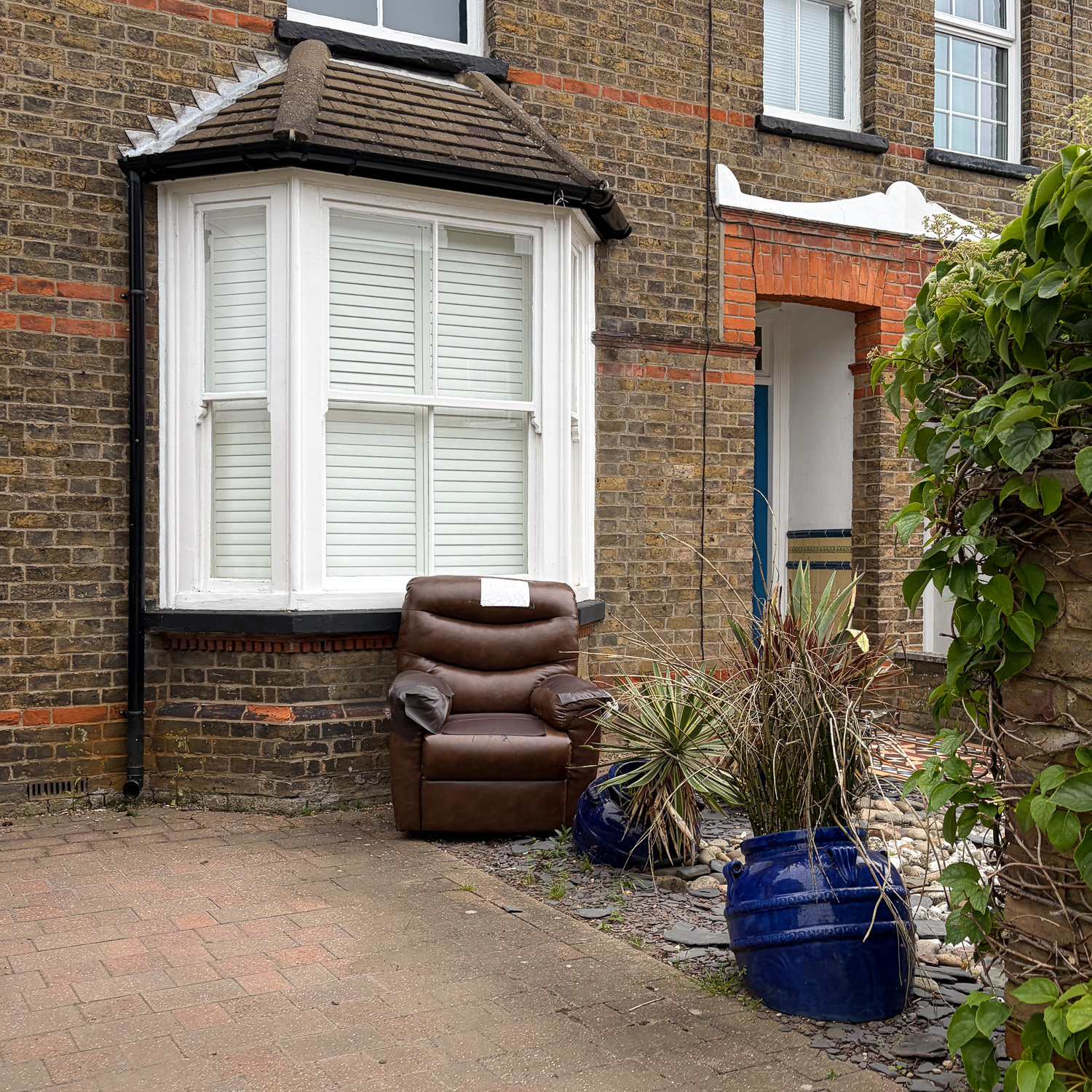 Photograph of a large brown leather armchair outside the bay window of a house, with a sign saying 'FREE' on it, and two blue pots with spiky plants growing out of them in front, at angles in the ground.